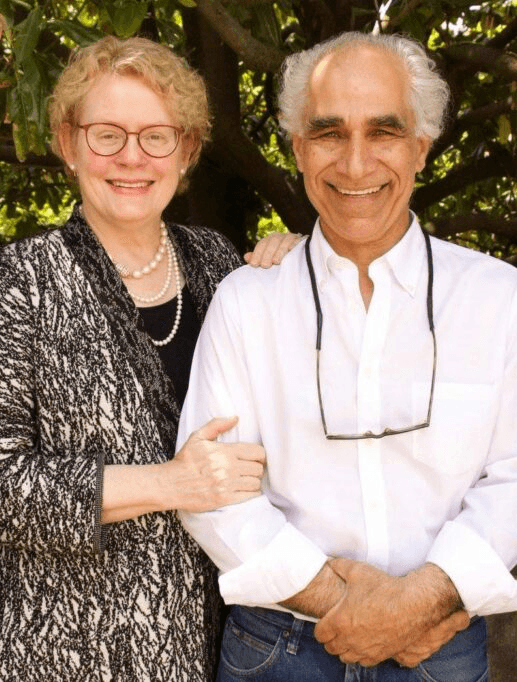 A smiling woman and man pose together in front of lush greenery.