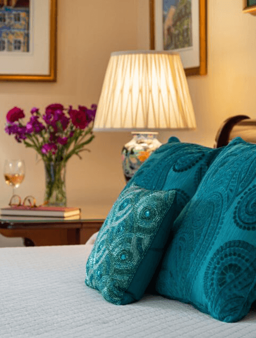 A cozy bedroom scene featuring turquoise pillows, a table lamp, a vase of flowers, and a glass of wine.