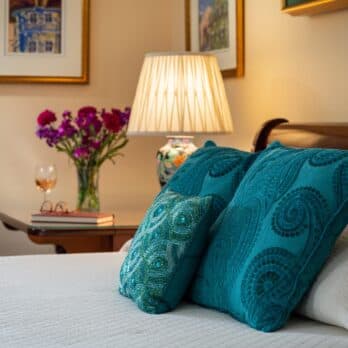 A cozy bedroom scene featuring teal decorative pillows, a bedside lamp, a vase of flowers, and a glass of rose wine.