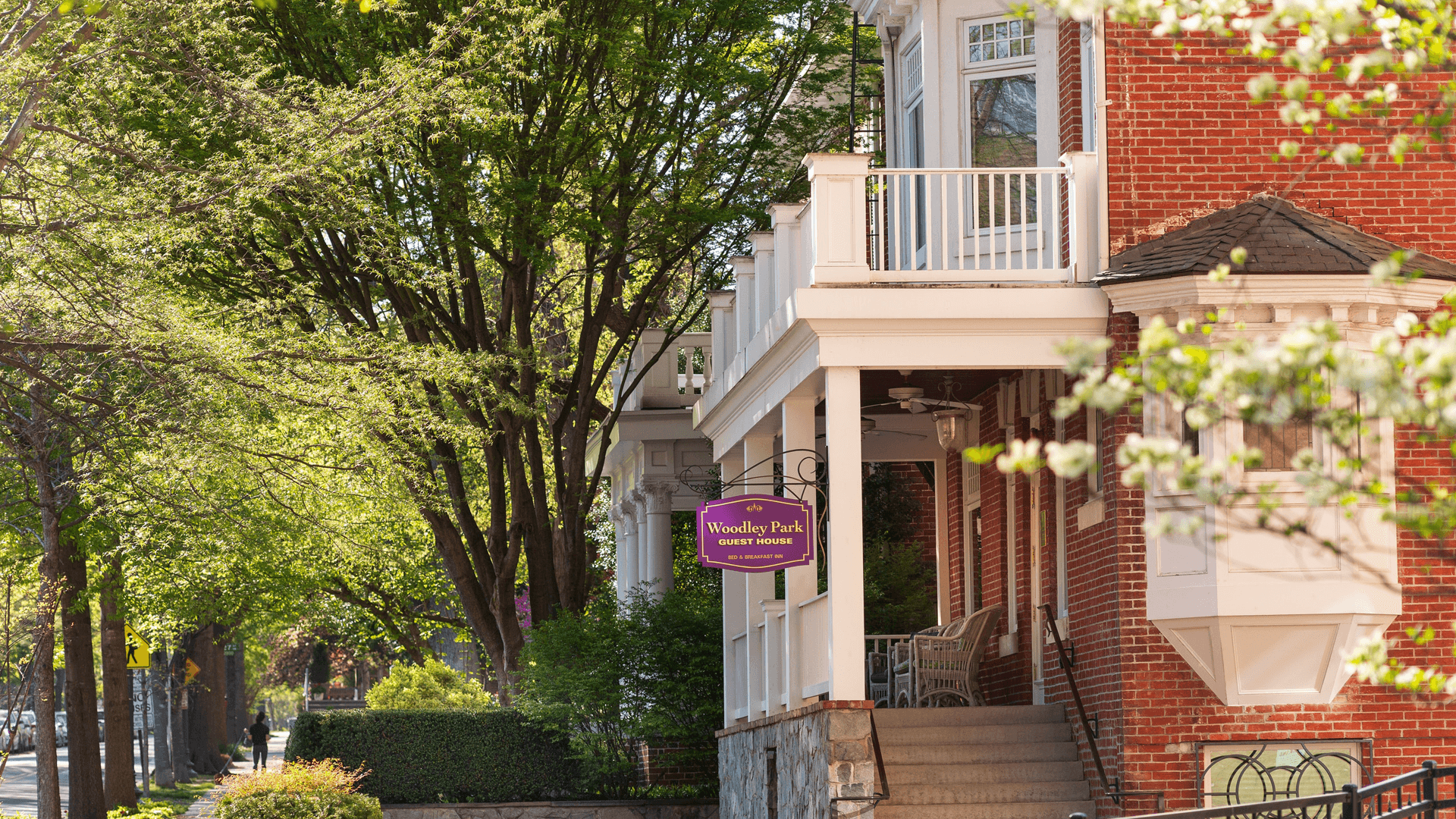 View of a red brick guest house with a sign reading "Woodley Park Guest House," surrounded by lush green trees.