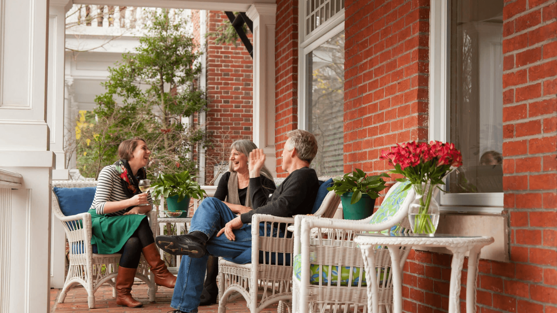 Three people enjoy a cheerful conversation on a porch surrounded by plants and flowers.