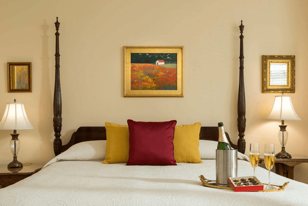 A cozy bedroom featuring a decorated bed with colorful pillows, framed artwork, and a tray with champagne and glasses.