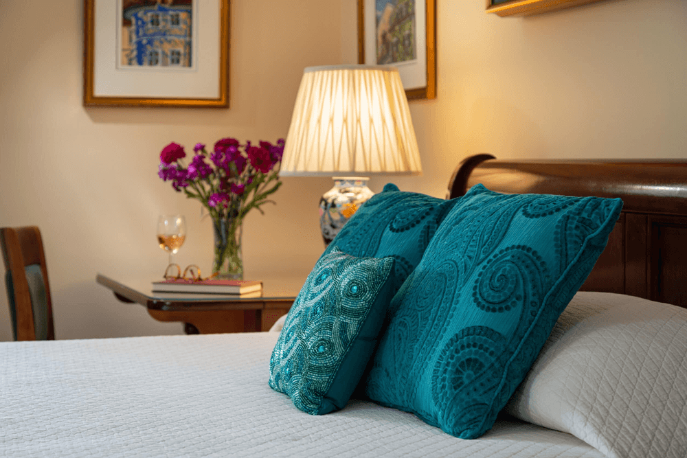 A cozy bedroom scene featuring decorative teal pillows, a bedside lamp, a vase of flowers, and a glass of wine.
