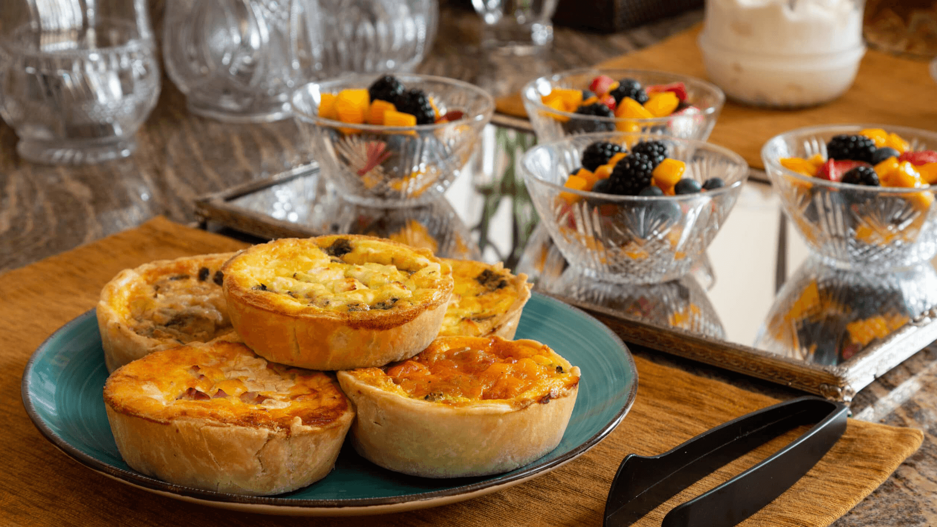 A plate of assorted savory quiches beside bowls of fresh fruit salad on a table.